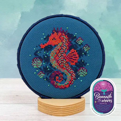 Displayed round cross stitch of a seahorse floating among fish and ocean bubbles stitched on a blue linen background.