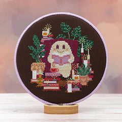 Displayed cross stitch of a ghost curled up in a chair, reading among stacks of books, glowing candles, leafy plants, and a steaming cup of warm brew on dark brown linen background.