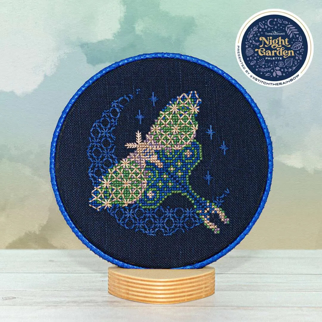 Displayed round cross stitch of a moth in flight in front of a glowing crescent moon.