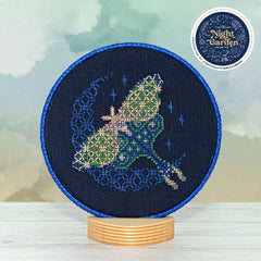 Displayed round cross stitch of a moth in flight in front of a glowing crescent moon.