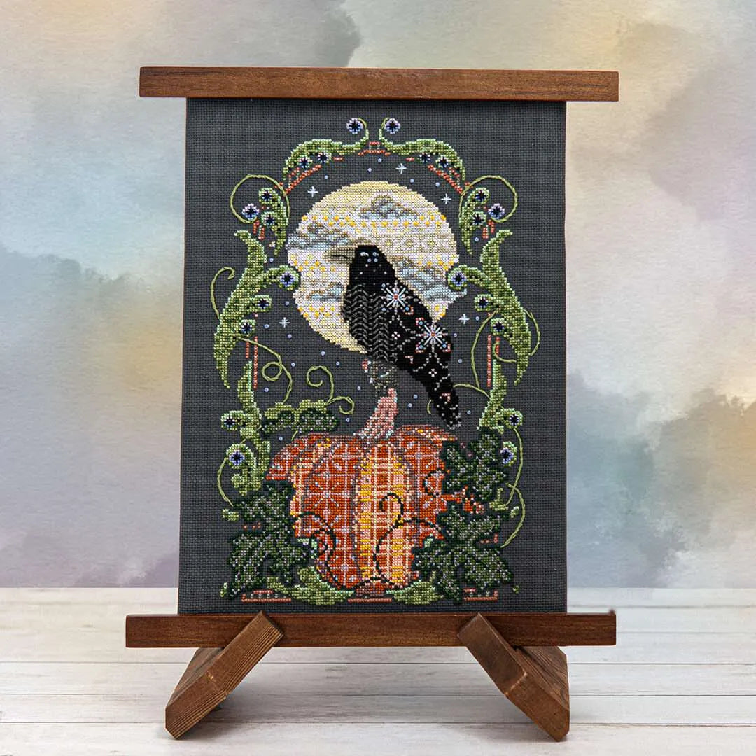 Displayed cross stitch of a raven perched atop a glowing pumpkin beneath a full harvest moon, surrounded by drifting clouds and curling foliage on a black linen background.