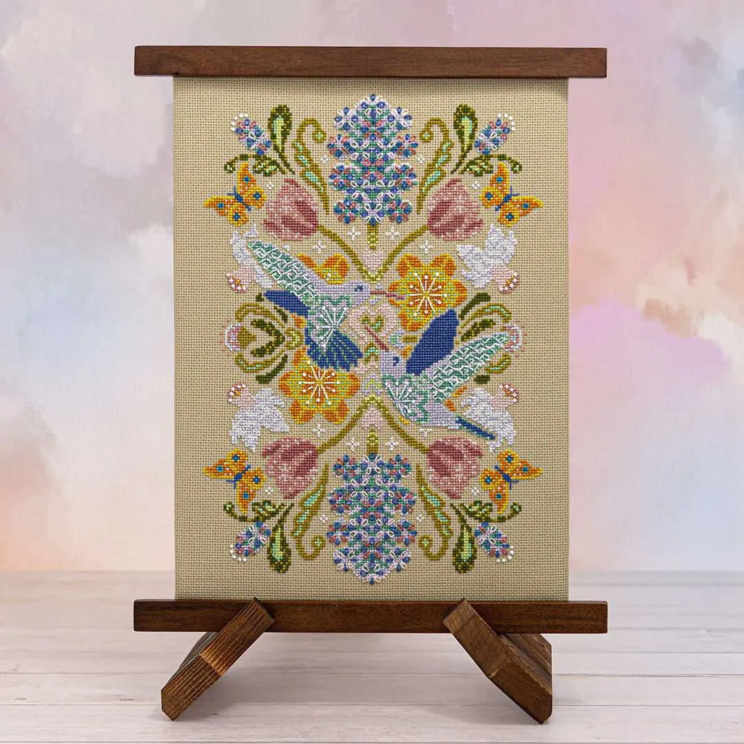 Displayed cross stitch of a mandala filled with hyacinths, daffodils, tulips, and alliums, accented by fluttering butterflies and two hummingbirds at the center.