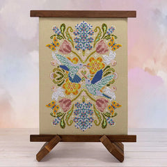 Displayed cross stitch of a mandala filled with hyacinths, daffodils, tulips, and alliums, accented by fluttering butterflies and two hummingbirds at the center.