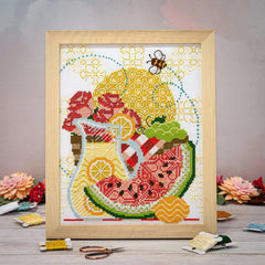 A cross stitch of bees, sunshine, lemonade, juicy watermelon, flowers, and a picnic basket in a wooden frame.