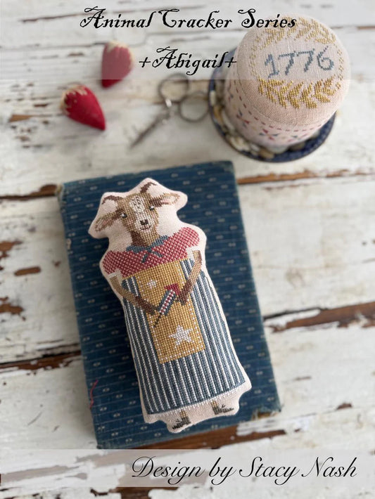 A cross stitch pillow of a farm goat dressed in a patterned dress and apron, and a pincushion drum with the year, "2026" stitched on top.