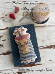 A cross stitch pillow of a farm goat dressed in a patterned dress and apron, and a pincushion drum with the year, "2026" stitched on top.