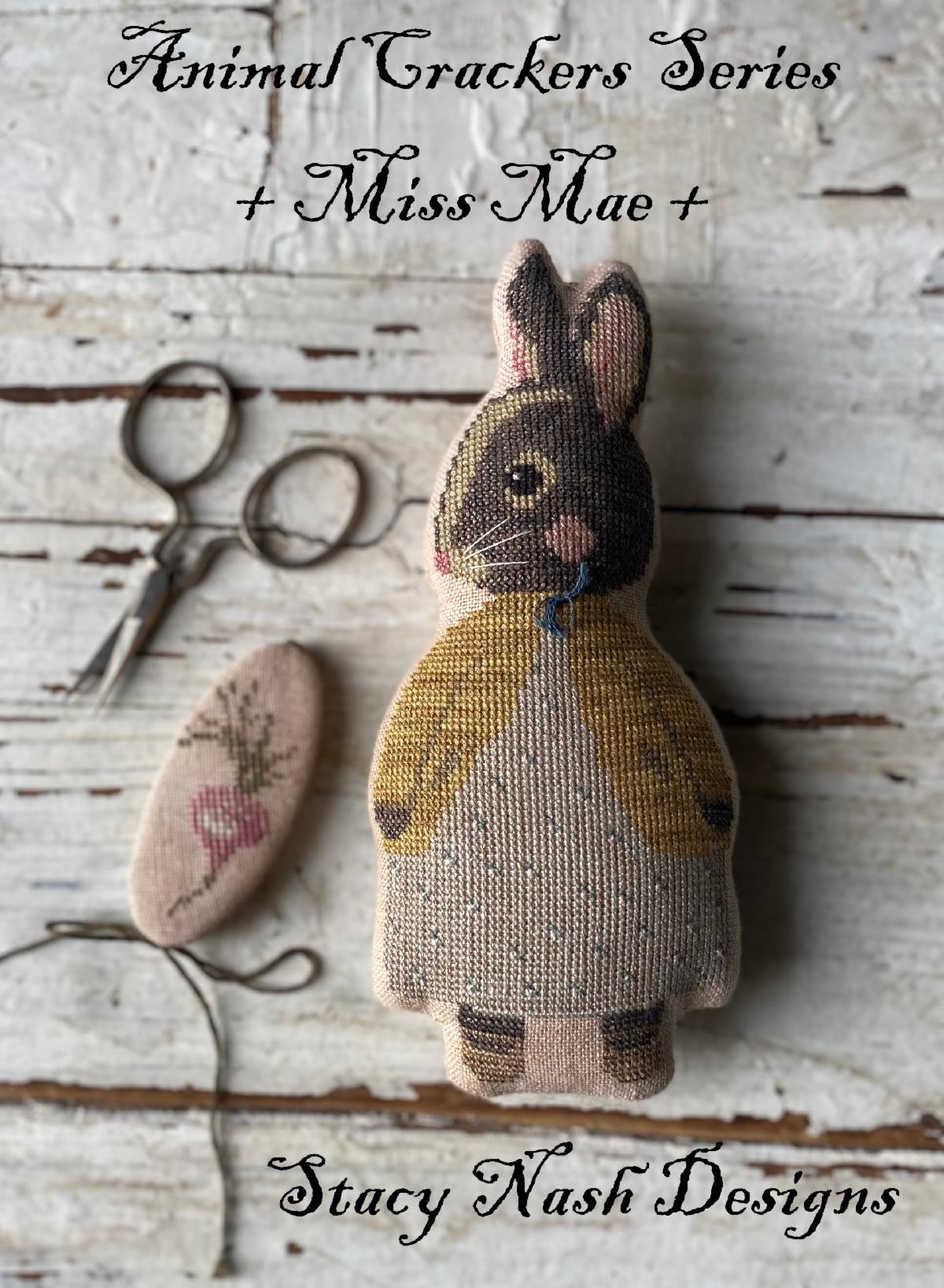 A cross stitch pillow of a bunny in a dress and cardigan next to a tiny beet‑stitched needlebook.