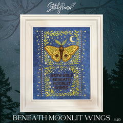 Framed cross stitch of a golden moth framed by leafy vines, stars, and a crescent moon on a deep blue background, with the phrase “Grow Bold Beneath Moonlit Wings” centered at the bottom.