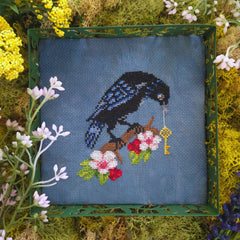 Cross stitch of a black-and-blue crow holding a gold key while perched on a branch with red and pink flowers, set against a dark blue background.