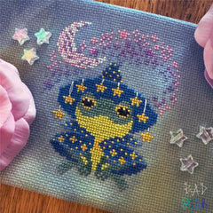 Cross‑stitch of a small blue-and-yellow frog in a starry wizard outfit under a crescent moon, surrounded by pink blossoms and decorative star accents.