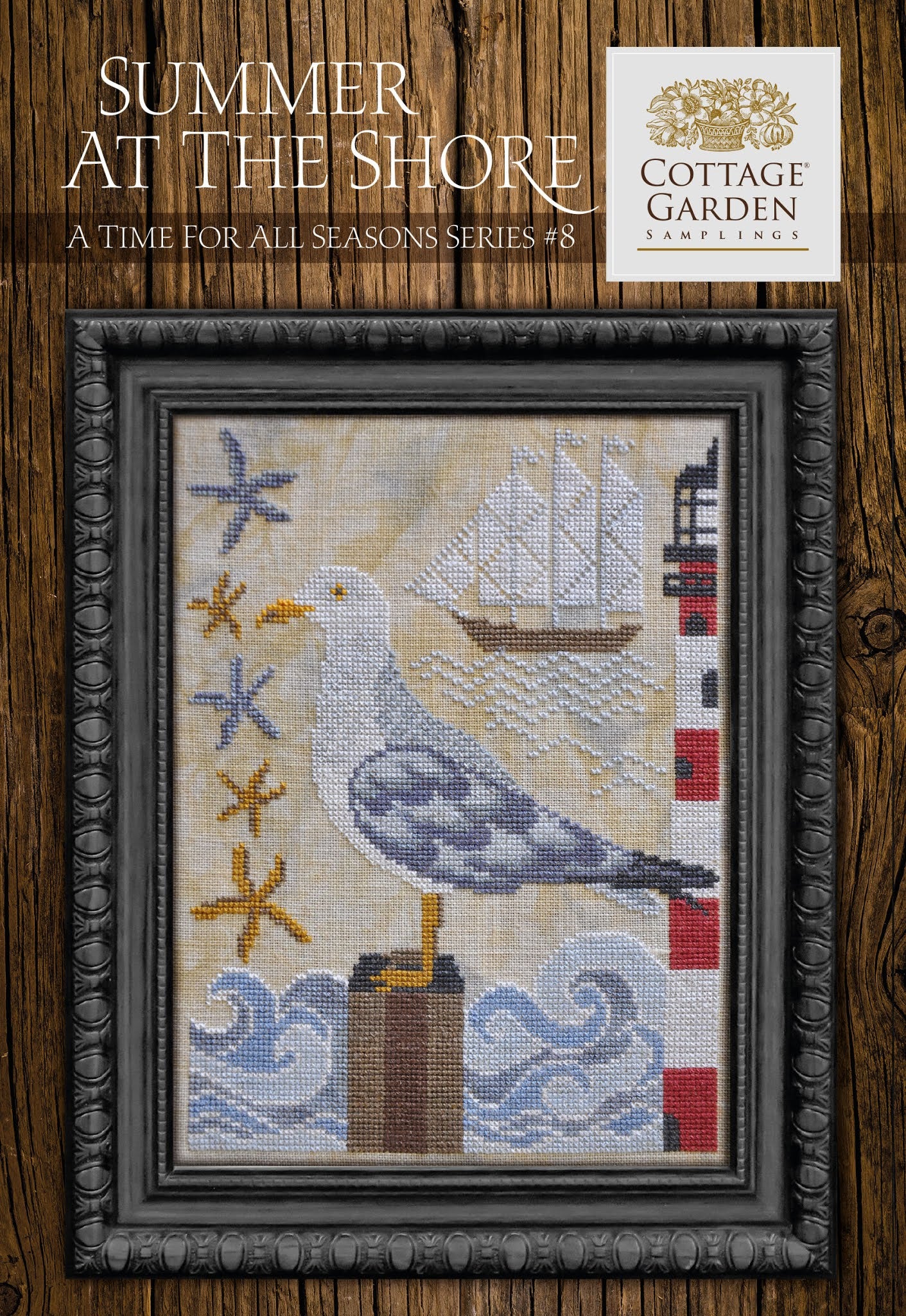 Framed cross-stitch sampler featuring a seaside scene with a seagull on a post, a lighthouse, a sailboat, waves, and scattered starfish on a neutral background.