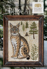 Framed cross stitch of a bobcat seated in a snowy woodland scene with evergreen trees and a small cabin.