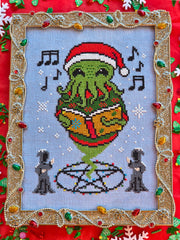 Cross stitch of a green tentacled Cthulhu creature wearing a Santa hat and singing from a book, surrounded by musical notes and snowflakes, with two robed figures below and framed in a gold tinsel‑like border decorated with colorful lights.