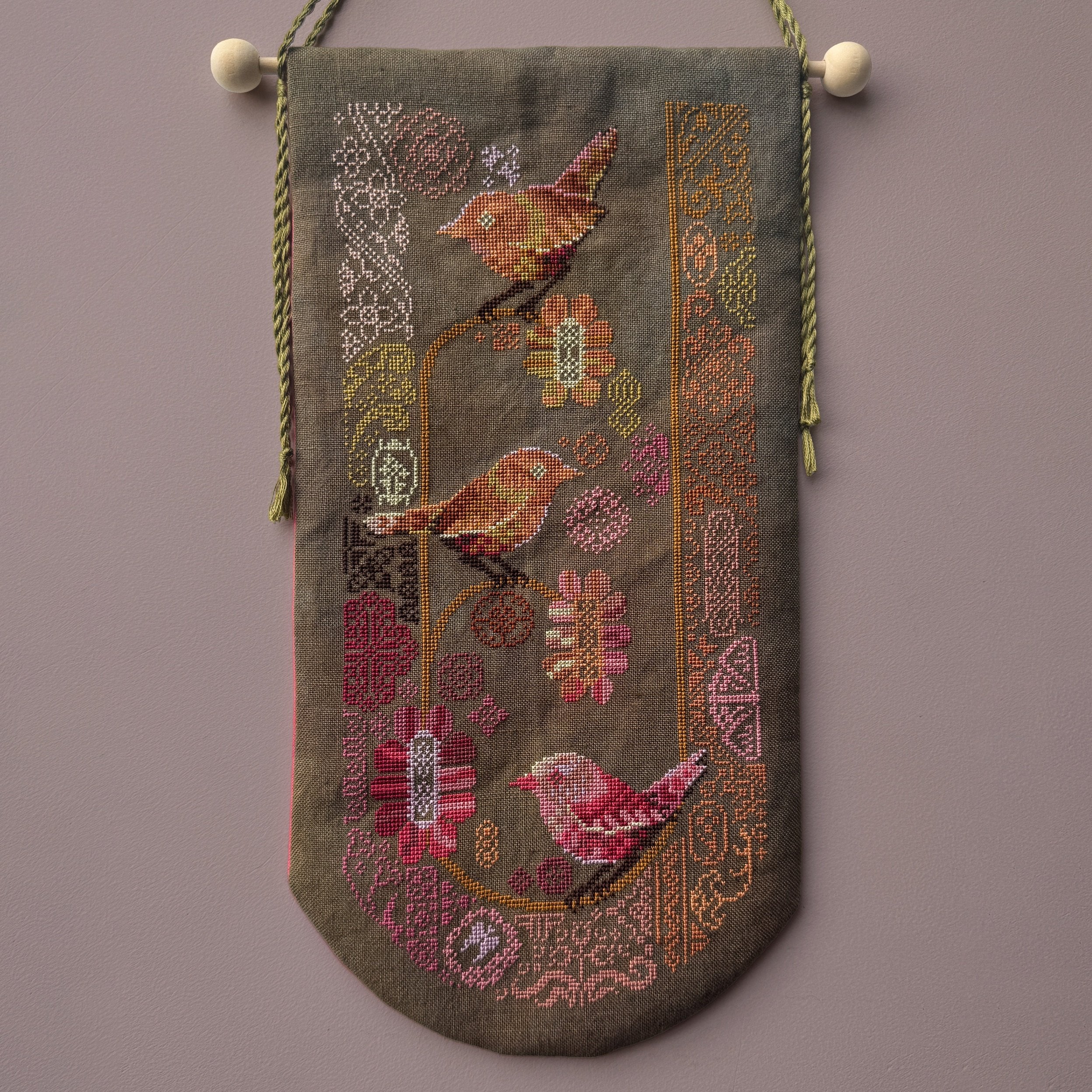 Cross stitched wall hanging featuring stylized birds perched among pink and orange flowers on a dark fabric background.
