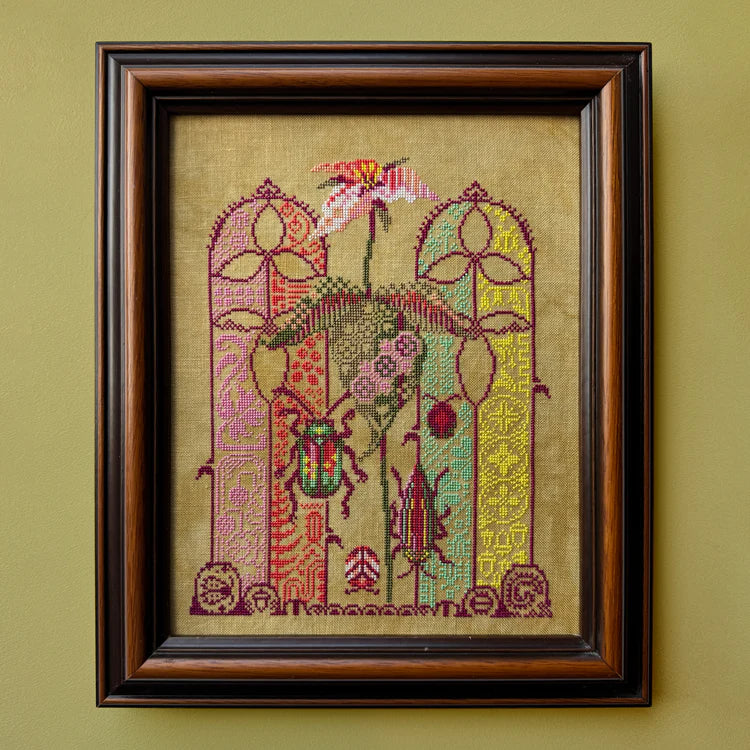 Framed cross‑stitch artwork depicting a white and pink trillium flower on a forest floor, surrounded by small beetles and intricate, colorful woodland motifs