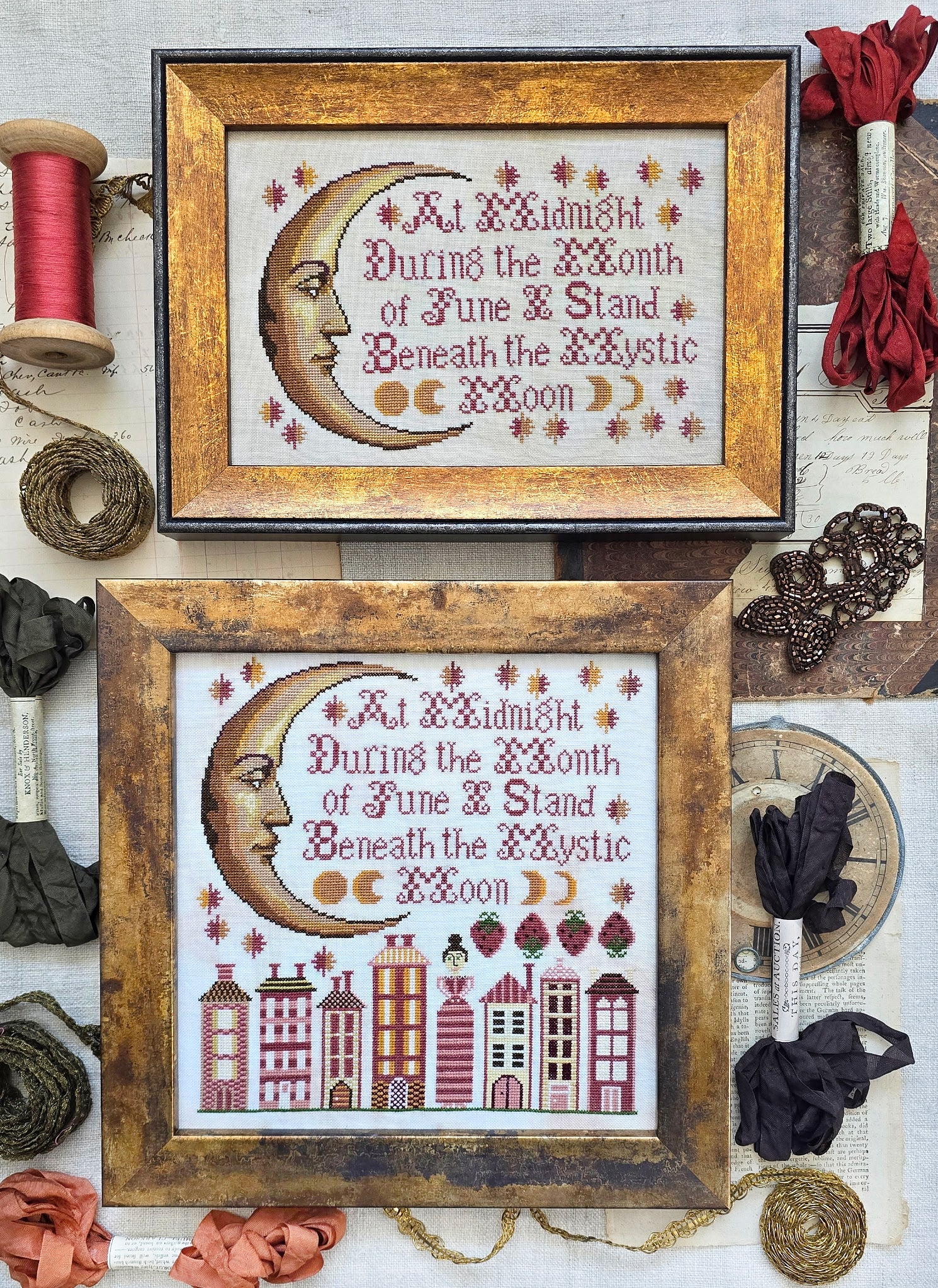 Framed cross-stitch samplers featuring a crescent moon face with decorative stars and stitched verse, with one design above a row of stylized houses, displayed with vintage sewing notions and ribbon.