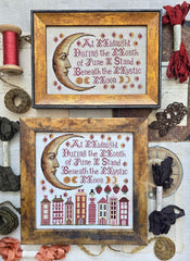 Framed cross-stitch samplers featuring a crescent moon face with decorative stars and stitched verse, with one design above a row of stylized houses, displayed with vintage sewing notions and ribbon.