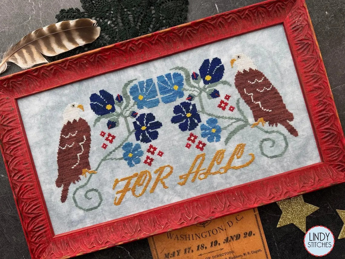 Cross stitch set at an angle of two bald eagles flanking a floral motif in blue and red, with the words “For All” stitched in mustard yellow, framed in a red carved frame.