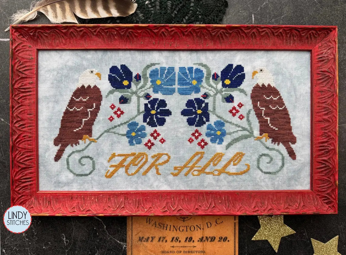 Cross stitch of two bald eagles flanking a floral motif in blue and red, with the words “For All” stitched in mustard yellow, framed in a red carved frame.