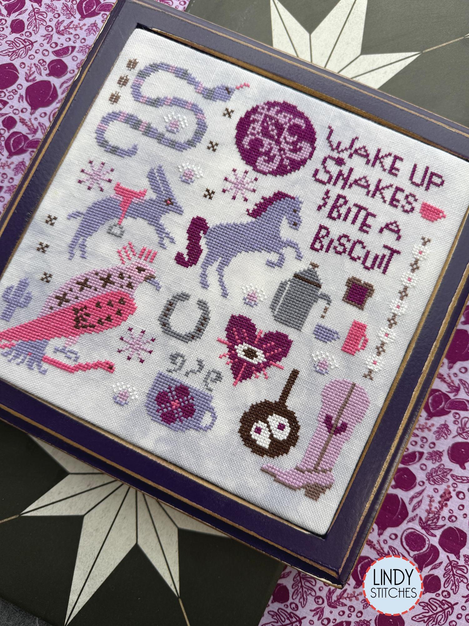 Right tilted angle view of framed cross stitch in pinks and purples with a horses, a rabbit, a snake, a bird, boots, a coffee pot, and the phrase “Wake up snakes, bite a biscuit.”