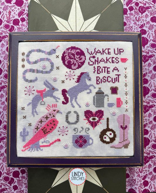 Framed cross stitch in pinks and purples with a horses, a rabbit, a snake, a bird, boots, a coffee pot, and the phrase “Wake up snakes, bite a biscuit.”