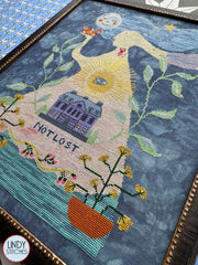 Side view of the cross‑stitch emphasizing the lower half, including the house, “Not Lost” text, potted plants, and water‑like stitching at the bottom.