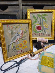 Two ornate gold-framed cross‑stitch pieces displayed on a wooden surface, one showing birds and flowers with ‘1910’ stitched below, the other depicting a pear and fig with ‘Aug 22 1910.’ Surrounding them are vintage sewing items including scissors, pink ribbon, a wooden bobbin, and a packet of rust‑proof needles.