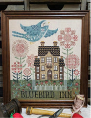 Cross stitch of a large blue bird flying above a tall brick house surrounded by tall stylized pink flowers, with “BLUEBIRD INN” stitched at the bottom.
