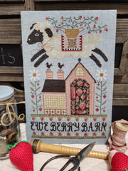 Cross stitch of a leaping sheep carrying a basket of flowers above a pink brick house filled with strawberries, framed by stitched florals and the words “EWE BERRY BARN" at the bottom.