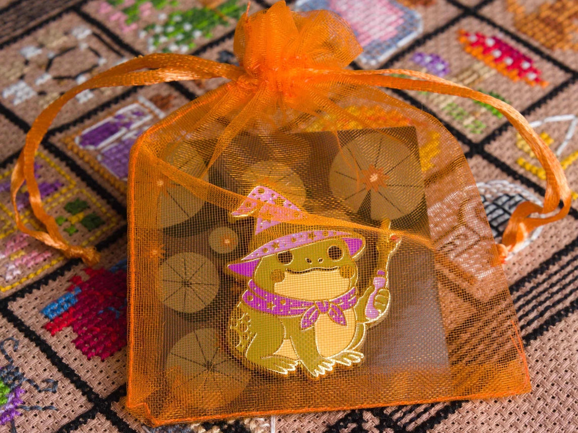 Enamel needle minder of a cute frog wizard in a purple hat and cape, shown in an orange mesh bag on a patterned backing card over a colorful cross stitched background.