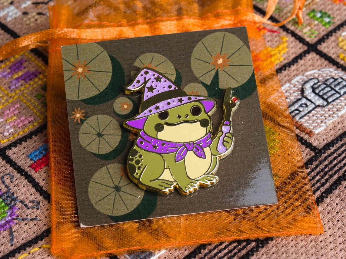 Enamel needle minder of a cute frog wizard in a purple hat and cape, shown on an orange mesh bag on a patterned backing card over a colorful cross stitched background.