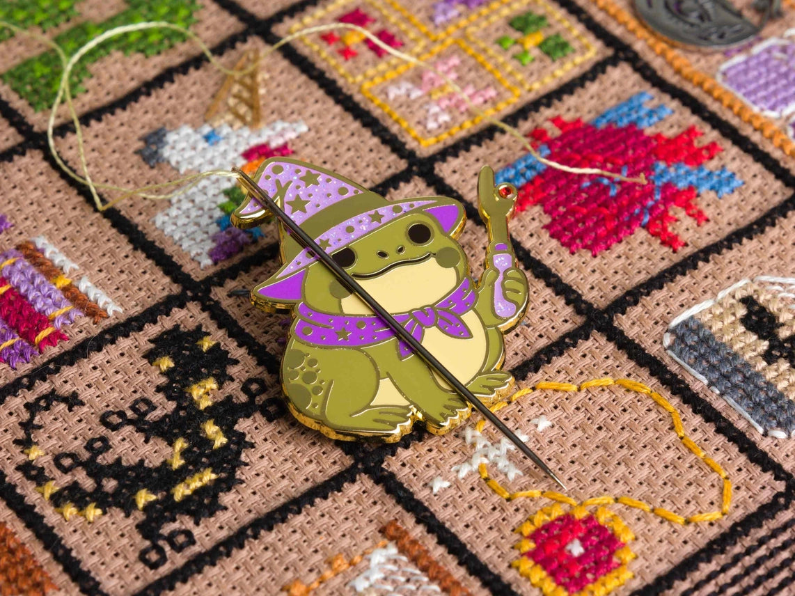 Enamel needle minder of a cute frog wizard in a purple hat and cape, shown holding a needle and thread on a patterned backing card over a colorful cross stitched background.