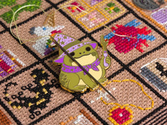 Enamel needle minder of a cute frog wizard in a purple hat and cape, shown holding a needle and thread on a patterned backing card over a colorful cross stitched background.