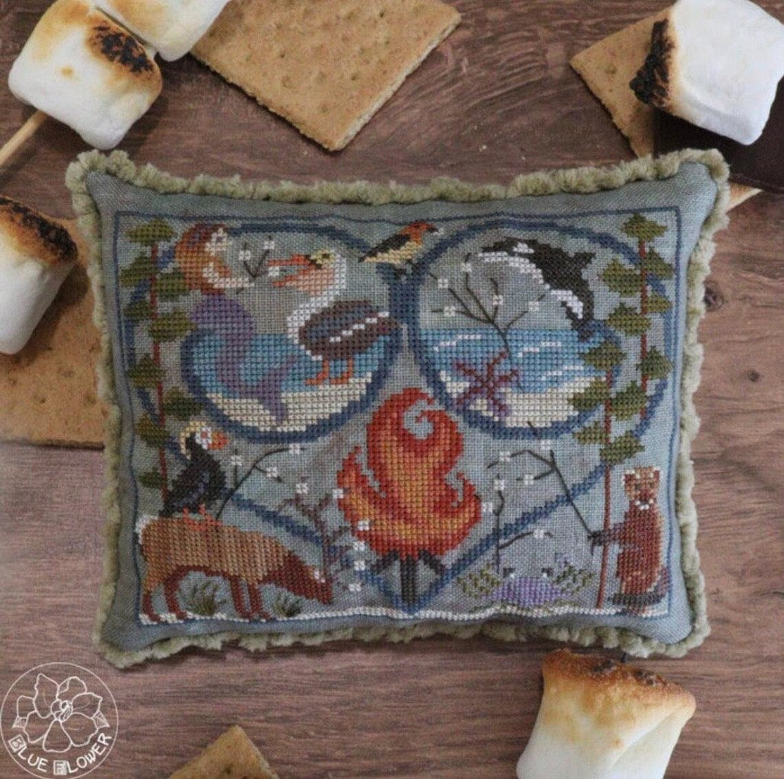 Cross stitched pillow showing a campfire scene with animals, birds, and seasonal motifs arranged in a heart frame.