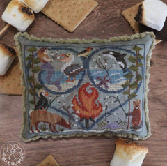 Cross stitched pillow showing a campfire scene with animals, birds, and seasonal motifs arranged in a heart frame.