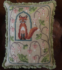 Cross stitch pillow featuring a red fox framed in an arch window, surrounded by curling vines and pink bleeding flower blossoms, with a green ruffled edge.