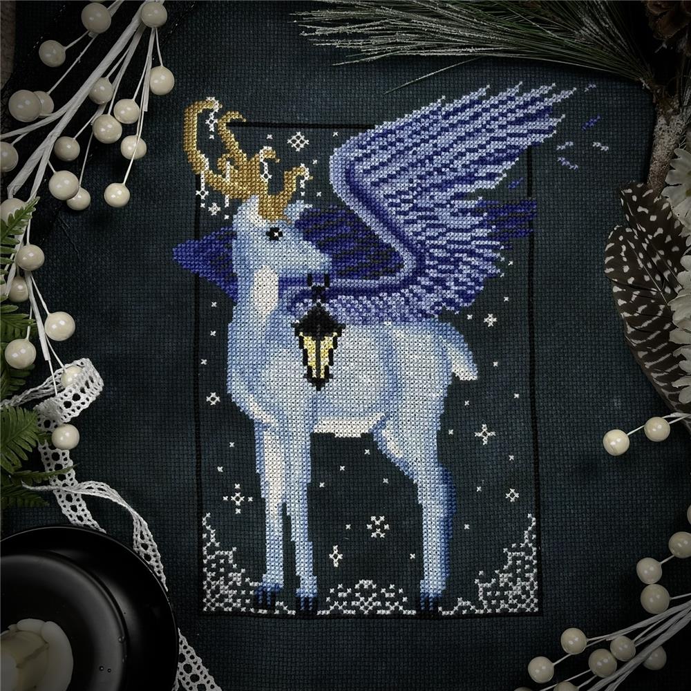 Cross stitch of a peryton winged deer with golden antlers holding a lantern, set against a dark snowy background and surrounded by winter foliage.