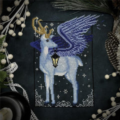 Cross stitch of a peryton winged deer with golden antlers holding a lantern, set against a dark snowy background and surrounded by winter foliage.