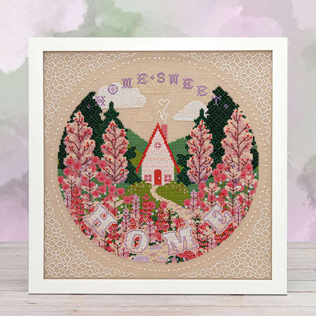 Framed cross stitch of a cottage among wildflowers, surrounded by tall trees and mountain peaks under clouds, with a winding stone path leading to the door and the words “Home Sweet Home” at the top.