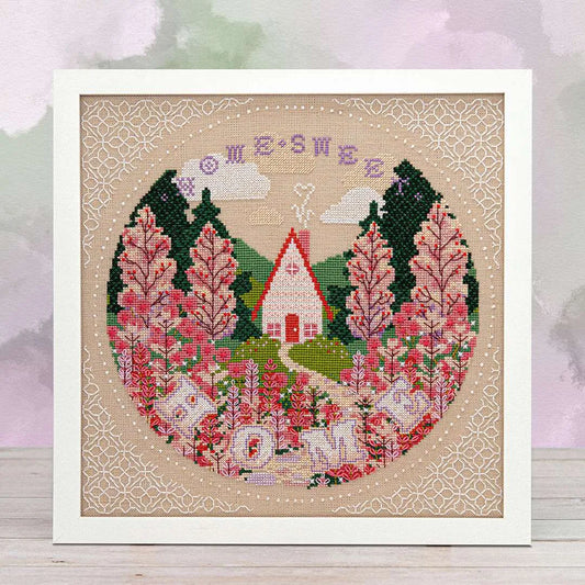Framed cross stitch of a cottage among wildflowers, surrounded by tall trees and mountain peaks under clouds, with a winding stone path leading to the door and the words “Home Sweet Home” at the top.