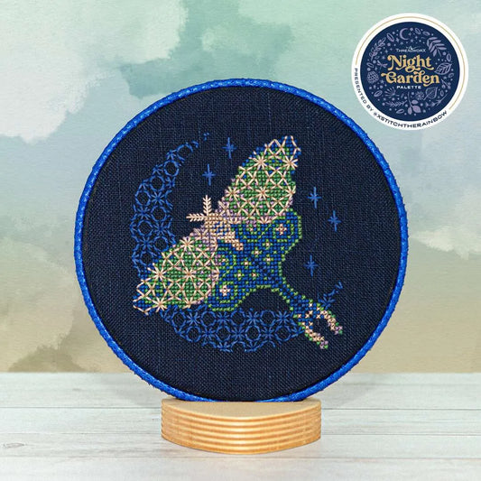 Displayed round cross stitch of a moth in flight in front of a glowing crescent moon.