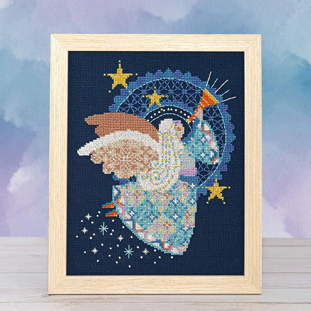 Wood framed cross stitch of an angel in flight, wearing a patchwork skirt, playing a horn, and surrounded by twinkling stars with white wings spread wide.