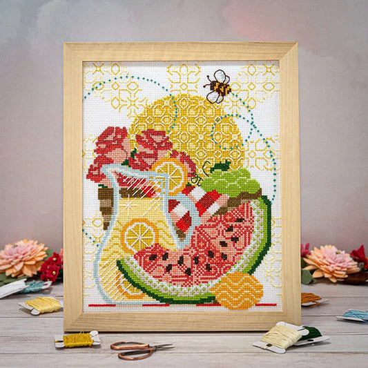 A cross stitch of bees, sunshine, lemonade, juicy watermelon, flowers, and a picnic basket in a wooden frame.