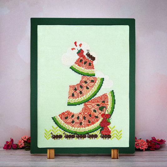 Framed cross stitch of ants hauling oversized slices of juicy watermelon, strawberries, and cherries on a green linen background.