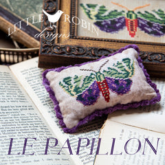 A cross stitch pillow of  butterfly with rich purple and green wings with purple fringe piping on the edges.