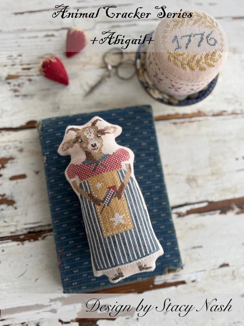 A cross stitch pillow of a farm goat dressed in a patterned dress and apron, and a pincushion drum with the year, "2026" stitched on top.
