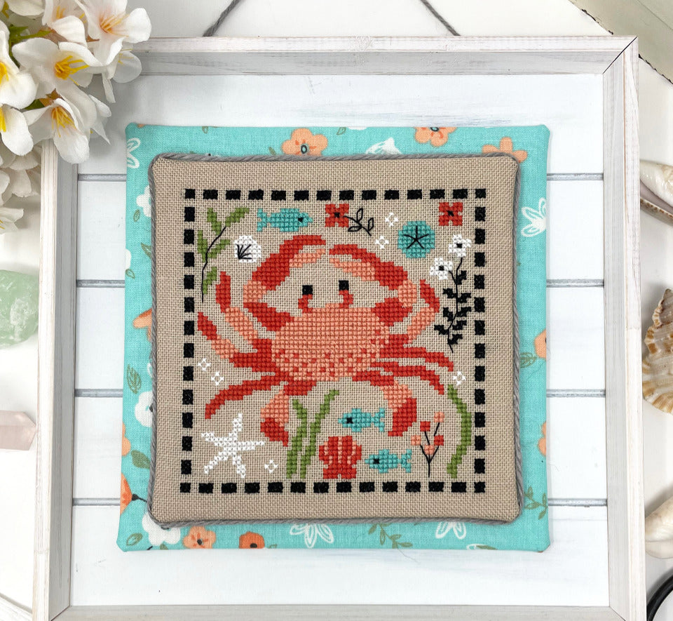 Framed cross stitch of a bright red crab surrounded by tiny sea creatures, plants, and geometric accents.
