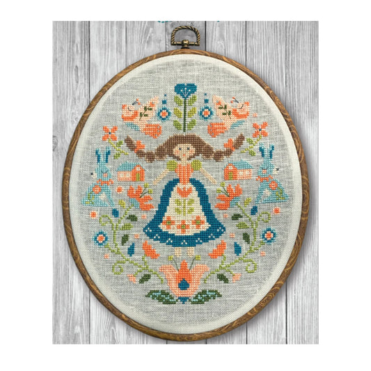 Oval framed cross stitch of a folk art sampler of a women with long braids in a dress surrounded by symmetrical flowers, leaves, birds, and hearts stitched in soft blues, corals, and green on a gray linen background.