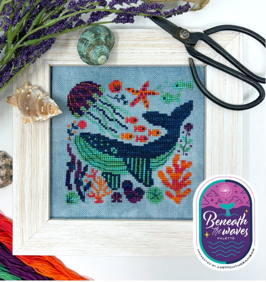 Wood framed cross stitch of a whale swimming underwater with jellyfish, coral, and swirling plants in rich blues and jewel tones. Styled with antique sewing scissors, sea shells and flowers on the edge of the frame.