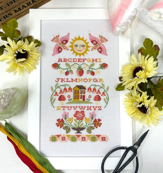 Framed cross stitch of a sampler with the alphabet, ripe strawberries, blooming flowers, a cozy little house, and a smiling sun.